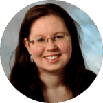 A woman with long brown hair, glasses, and earrings smiles at the camera. She is wearing a dark top and is against a blurred, light-colored background.