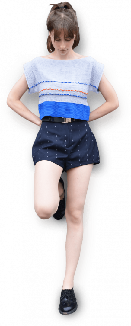 A person stands with one knee bent, wearing a color-blocked cropped top in shades of gray and blue, crafted from soft Merino wool. They pair it with high-waisted navy shorts adorned with white polka dots, looking down with hands on hips, and finish the look with sleek black shoes.