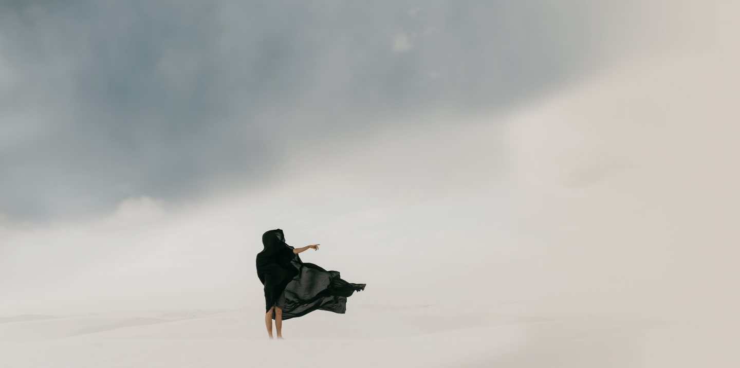 A person wearing a black cloak crafted from fine Merino wool stands on a vast, white sand dune under a cloudy sky. They face the horizon, with one arm outstretched, as the wind blows their cloak dramatically. The scene conveys a sense of solitude and vastness.