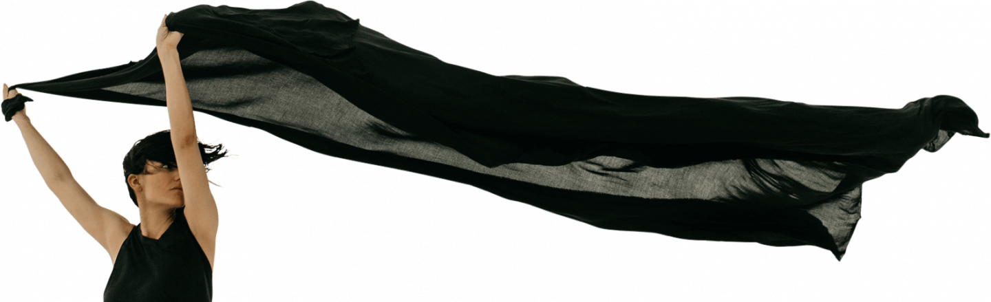 A person wearing a black sleeveless top energetically holds up a flowing merino wool fabric above their head, with the cloth trailing dramatically in the air behind them. The background is plain white, emphasizing the movement of the luxurious yarn.