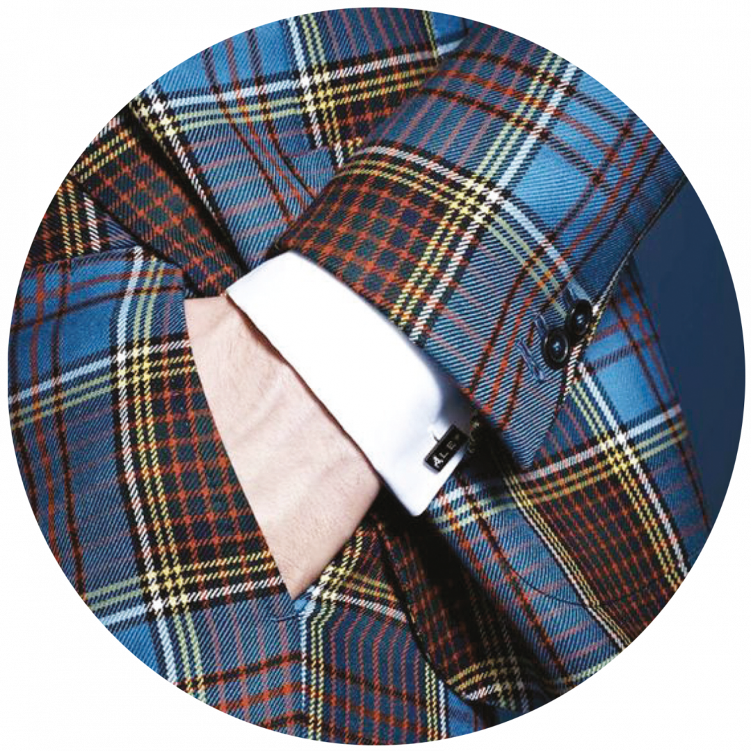 A person wearing a blue and brown tartan plaid suit crafted from fine wool with one hand in the pocket. The suit jacket features dark buttons, and the person is wearing a white shirt underneath with visible cufflinks.