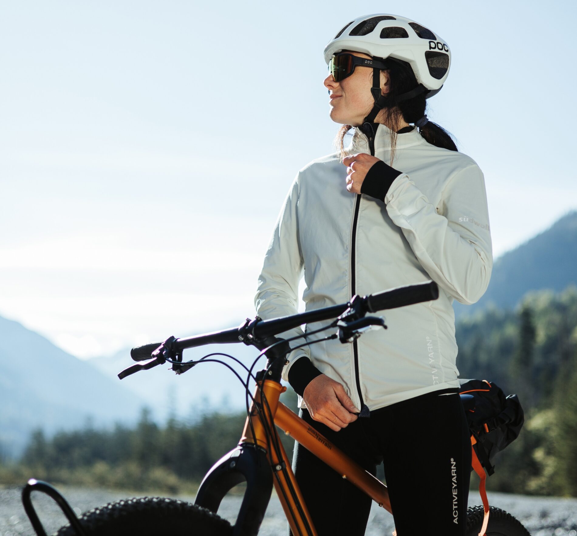 A woman wearing a white helmet, sunglasses, and a light jacket stands beside an orange mountain bike outdoors, looking into the distance with mountains and trees in the background.