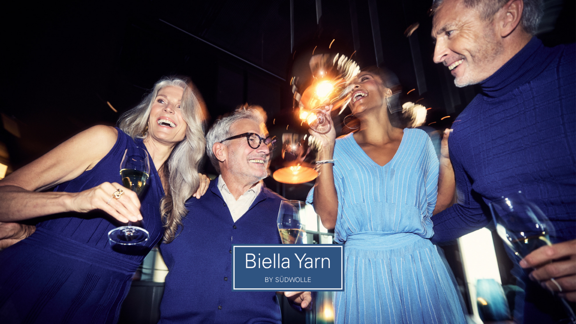 Four smiling adults in blue outfits holding drinks and sparklers celebrate together. The text “Biella Yarn by Südwolle” appears at the bottom center. The atmosphere is festive and joyful.