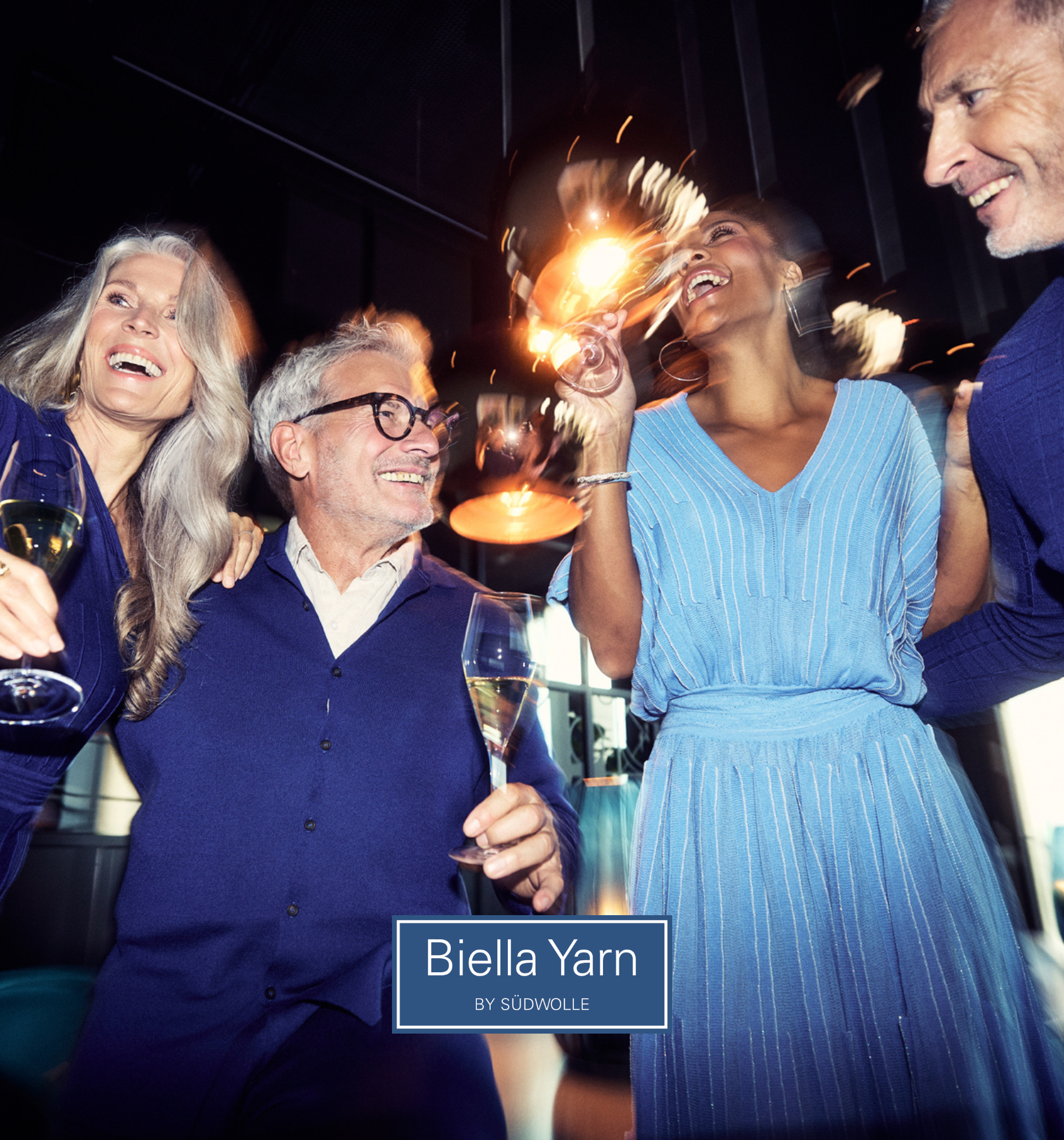 Four smiling adults dressed in blue hold glasses of champagne and sparklers, celebrating together indoors. The text Biella Yarn by Südwolle appears at the bottom of the image.