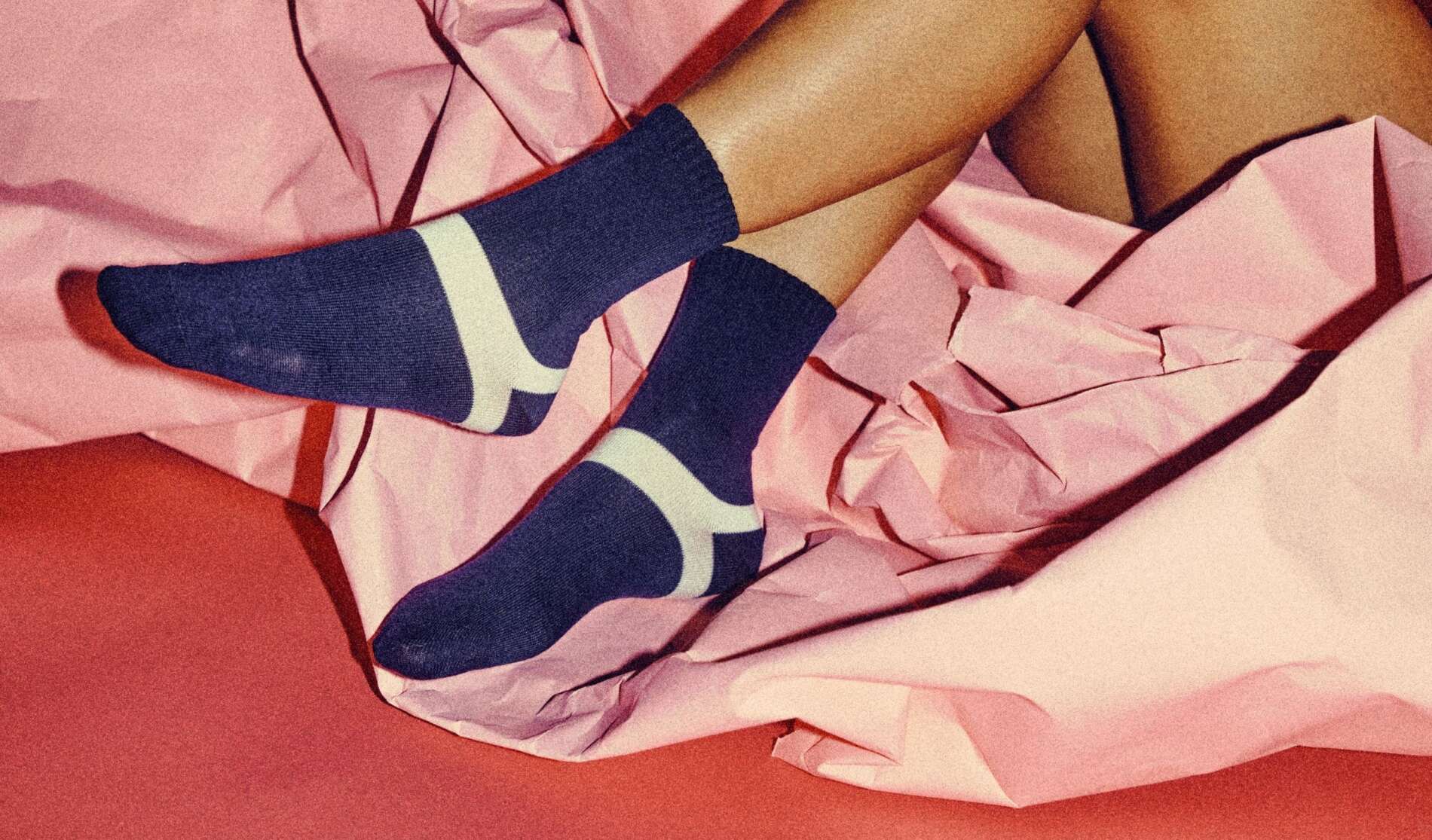 Person wearing dark blue socks with a white stripe lies on pink crumpled paper, their legs crossed and relaxed. The background is also pink, creating a playful and cozy atmosphere.