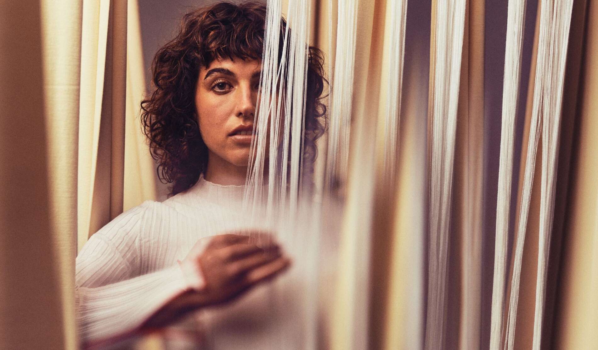 A person with curly hair and a white top looks through vertical threads, with one hand touching the threads. The focus is on their face, partially obscured by the strands.