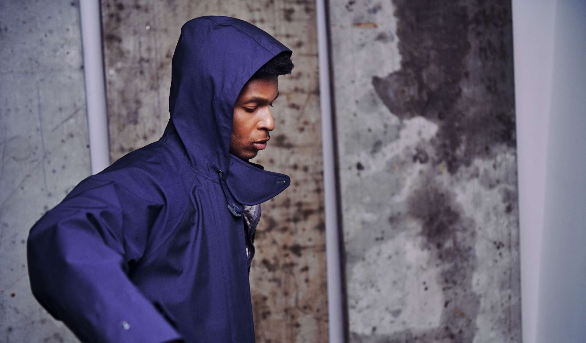 A person in a blue hooded jacket stands in front of a textured concrete wall, looking down with their face partially visible in profile.