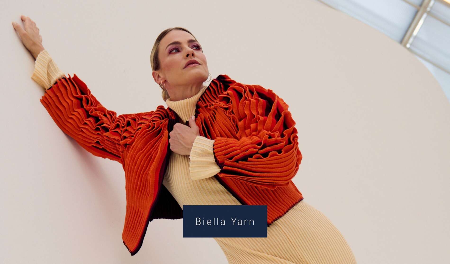 Woman in a pleated orange jacket and cream ribbed dress leans against a curved white wall. She looks upward, with one arm raised. Text reads Biella Yarn. Modern, stylish setting.