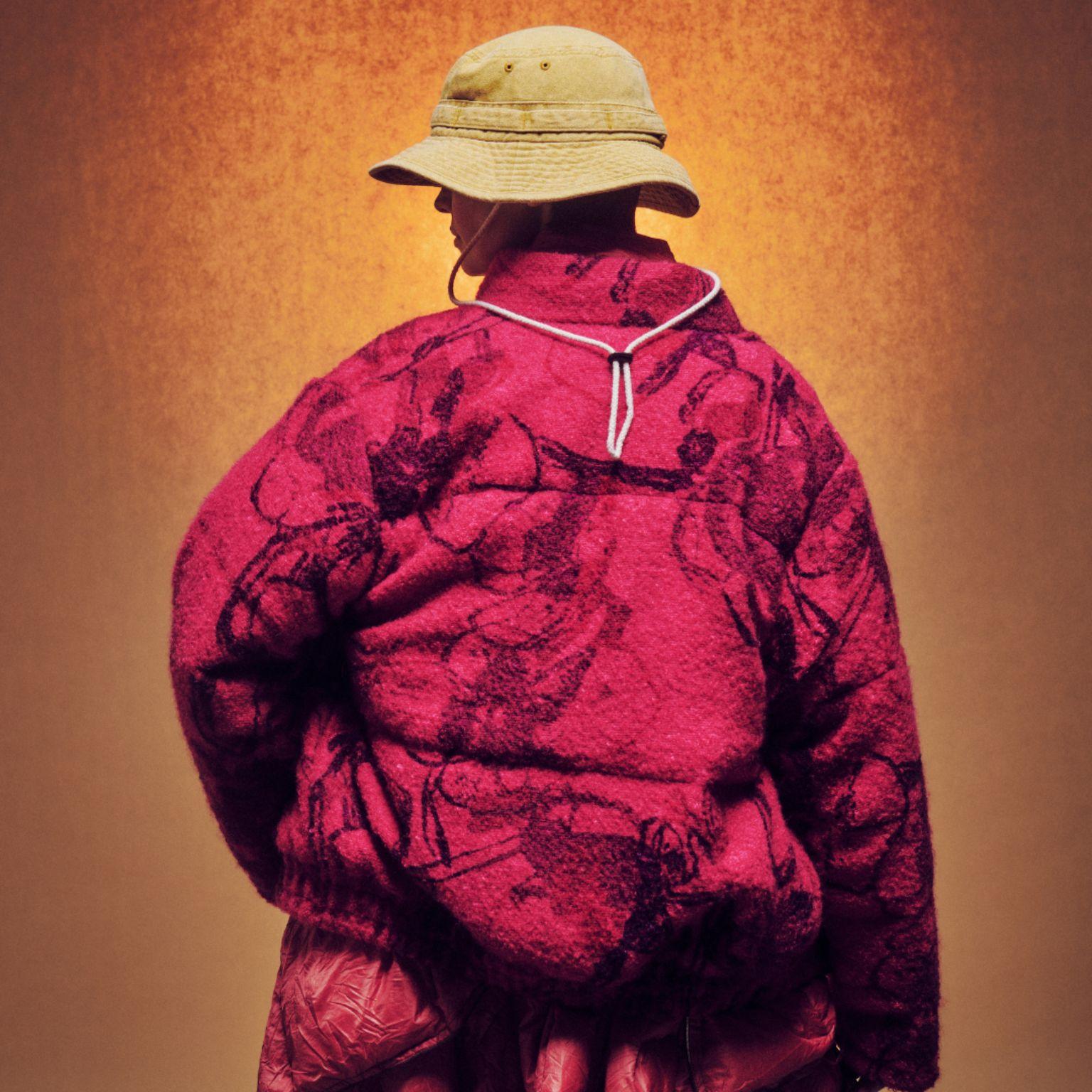 Person facing away, wearing a tan bucket hat and an oversized textured red jacket with dark floral patterns, against a warm orange-brown background.