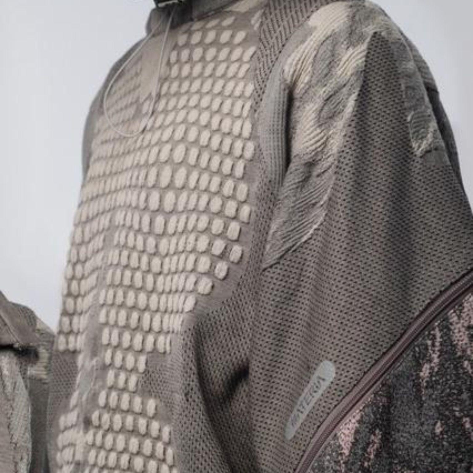 Close-up of a textured gray and beige sweater with mesh-like patterns, raised square shapes, and mixed knit materials. A small tag with text is visible on the right sleeve near a zipper detail.
