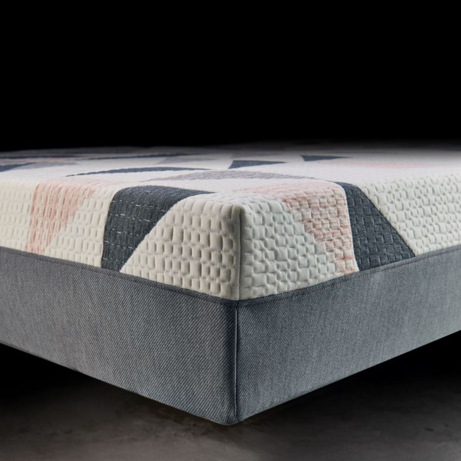 Close-up of a mattress corner showing a textured, patterned white and pastel fabric top with a gray fabric base, set against a dark background.