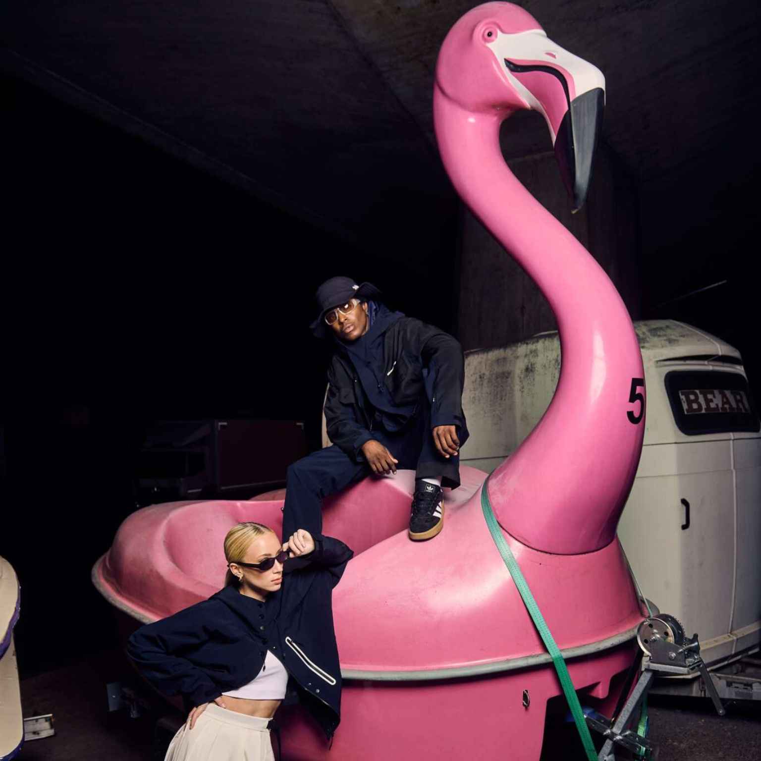 Two people pose with a large pink flamingo paddle boat. One sits on the flamingo, wearing dark clothing and sunglasses; the other stands in front, also in dark clothing and sunglasses, under a shadowy, industrial setting.