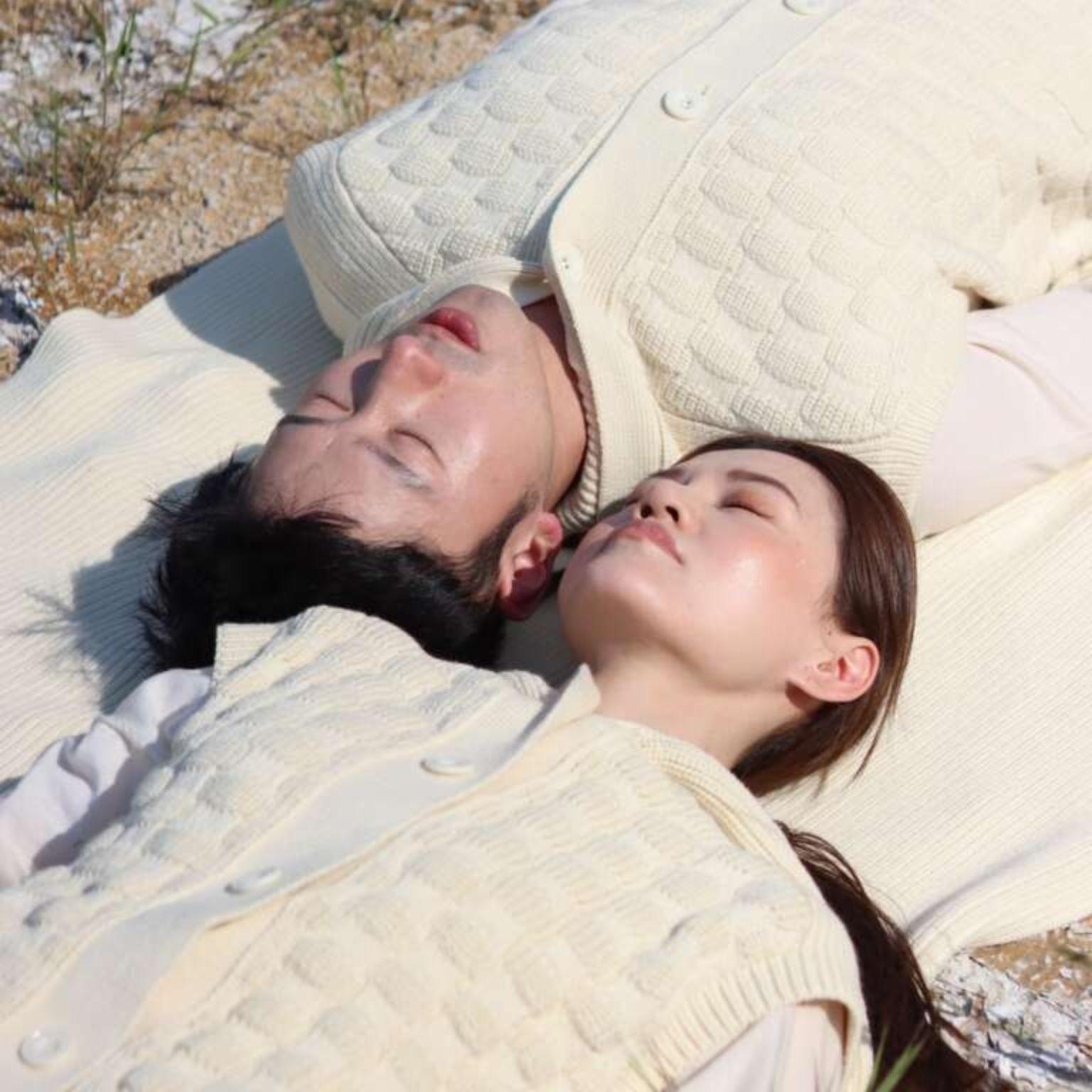 Two people wearing cream-colored textured vests lie on the ground with their eyes closed, facing up and appearing peaceful, with sunlight casting soft shadows on their faces. Sparse grass and sandy earth are visible around them.