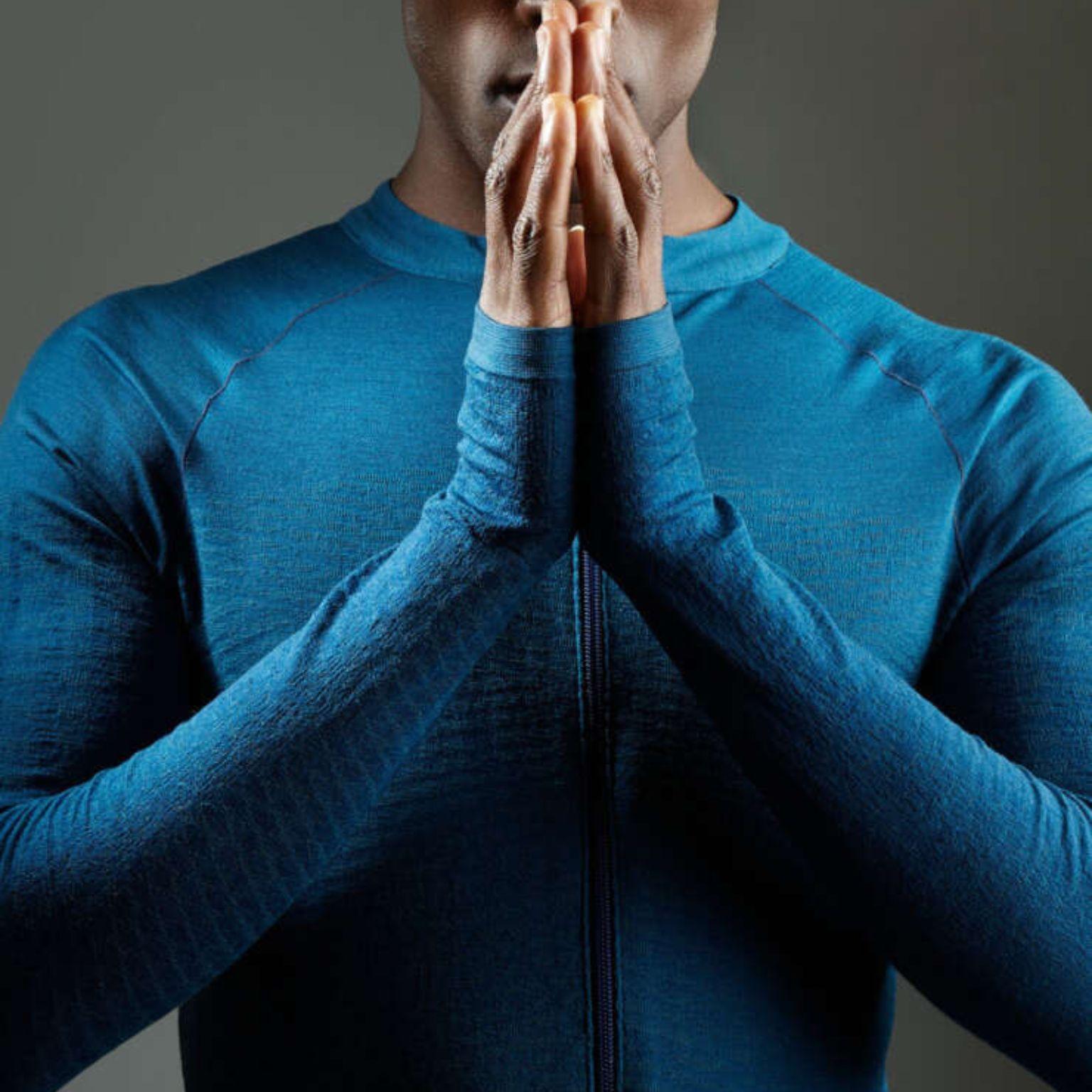 A person wearing a blue long-sleeve shirt holds their hands together in front of their face in a prayer or meditation pose, with a calm, neutral background.