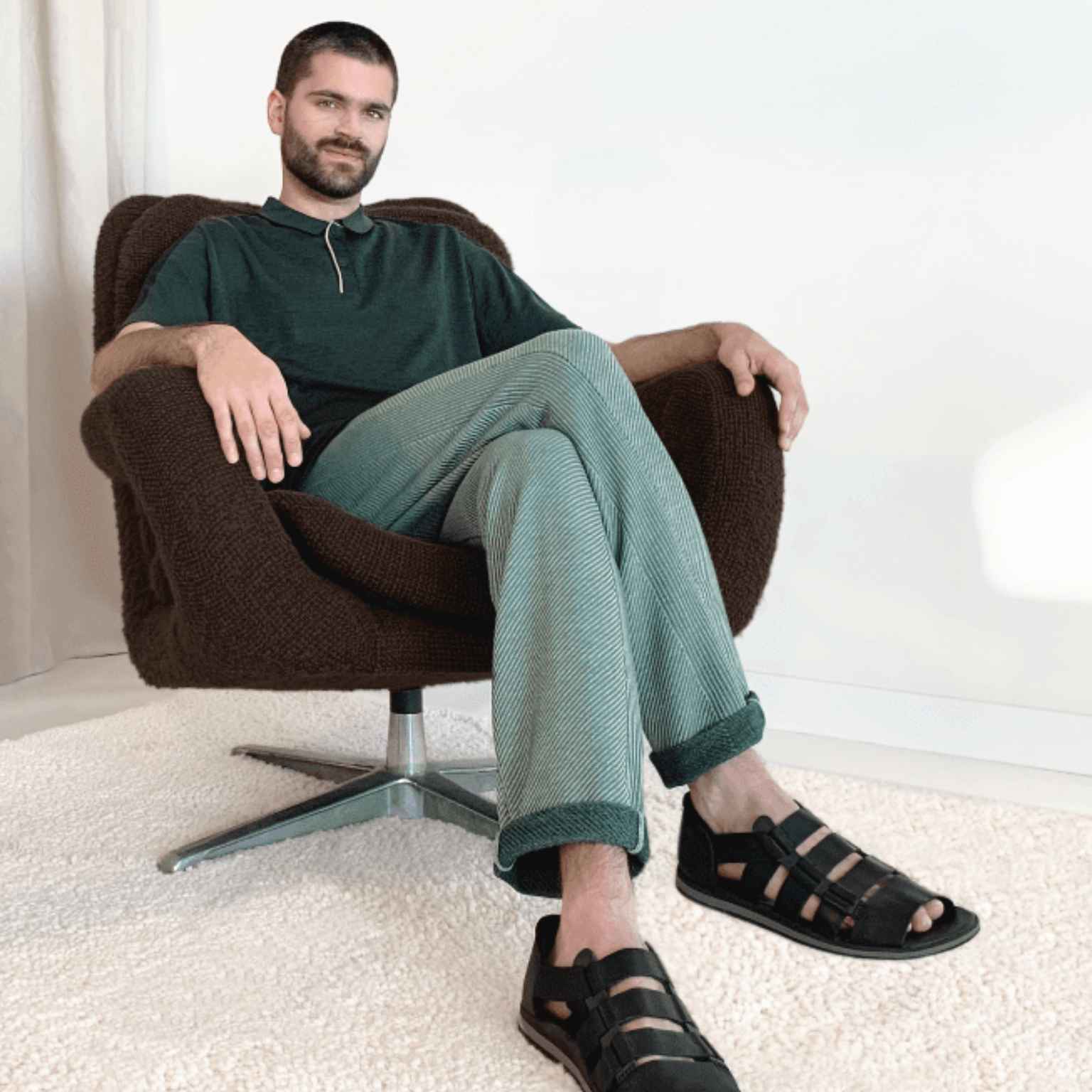 A man with a beard sits on a brown swivel chair, legs crossed, wearing a dark green shirt, light green pants, and black sandals, on a beige carpet in a minimalist room.