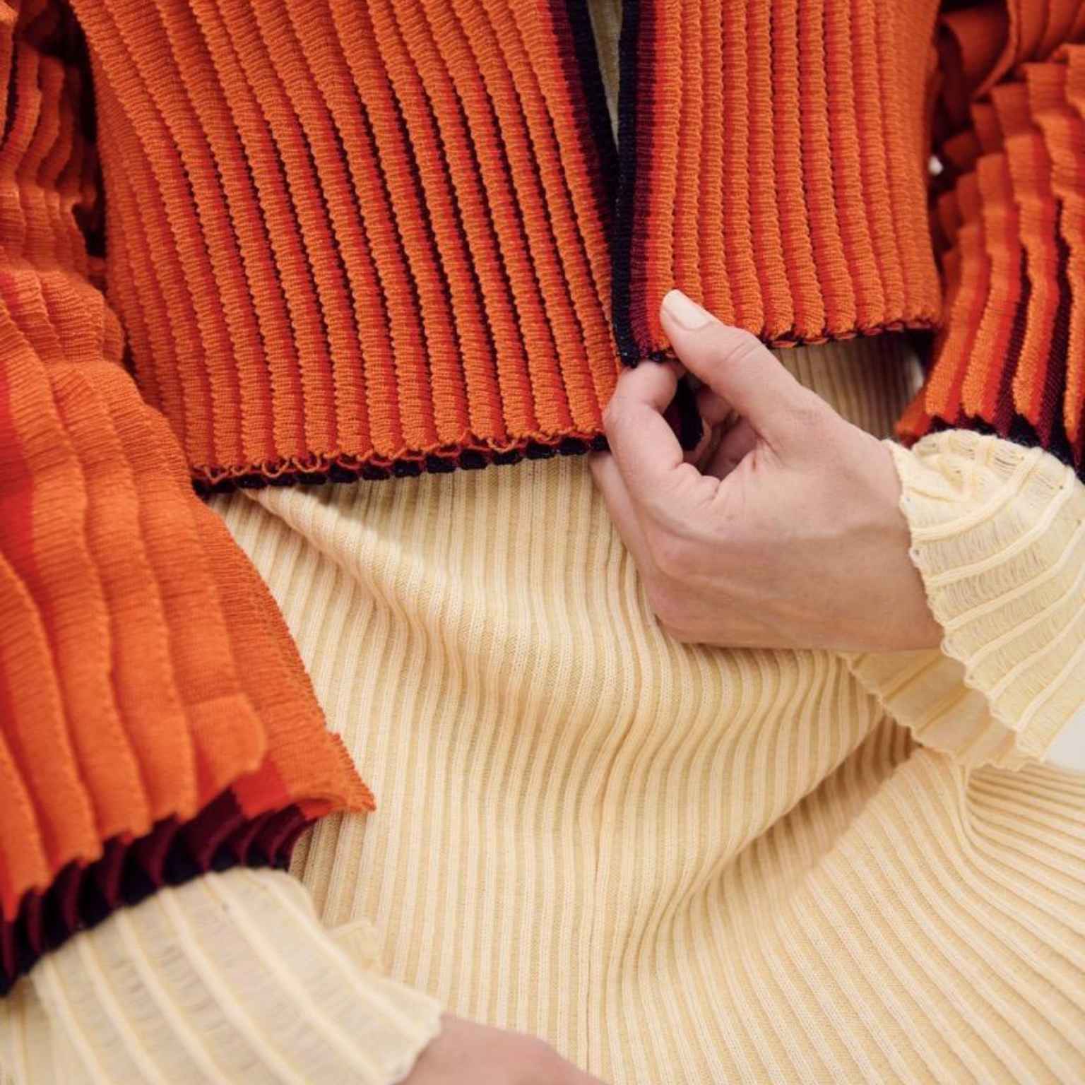 A person wearing a textured orange jacket over a cream ribbed dress is shown adjusting the front of the jacket with one hand. The image focuses on the clothings fabric details.