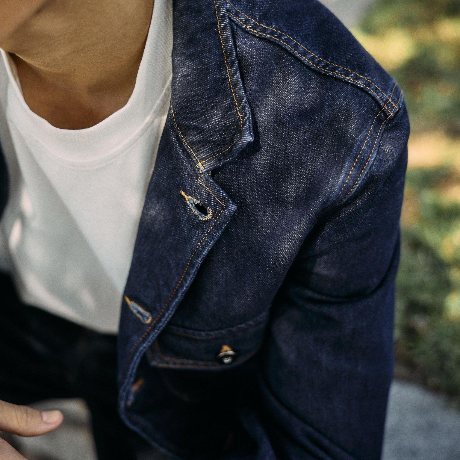 Close-up of a person wearing a white t-shirt and a dark blue denim jacket with tan stitching, seated outdoors on a sunny day. Only the lower face, neck, and clothing are visible.