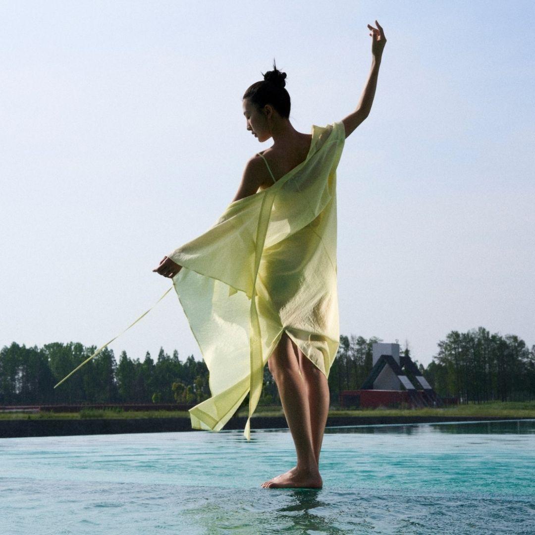 A woman in a flowing, sheer yellow dress stands gracefully on one foot by the water, arm raised, with trees and a building in the background under a clear sky.